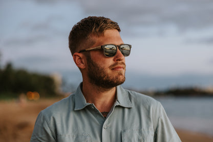 A person wearing Johnny Fly Splinter sunglasses featuring a modern minimal design with an acetate front, wooden arms, spring hinges, and nylon polarized lenses, enjoying a scenic beach view.