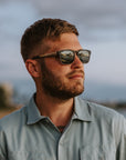 A person wearing Johnny Fly Splinter sunglasses featuring a modern minimal design with an acetate front, wooden arms, spring hinges, and nylon polarized lenses, enjoying a scenic beach view.